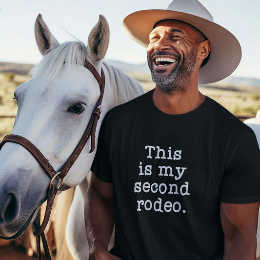 Laughing man standing next to a horse. He is wearing a black t-shirt with the funny phrase This is my second rodeo printed on the front.