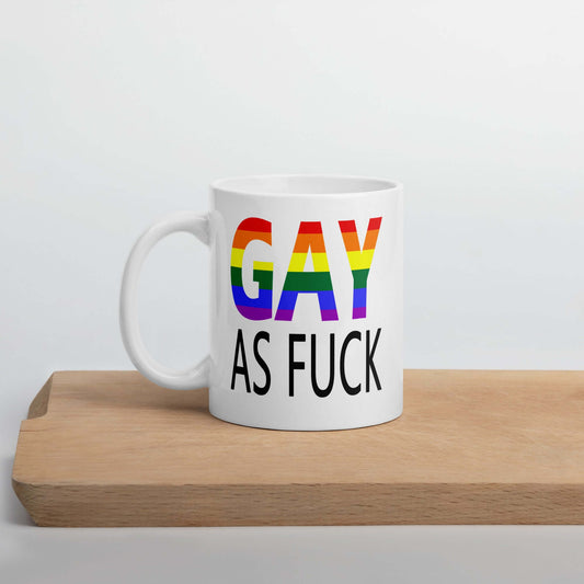 White ceramic mug with the words Gay as fuck printed on both sides. The word Gay is in rainbow stripe font.