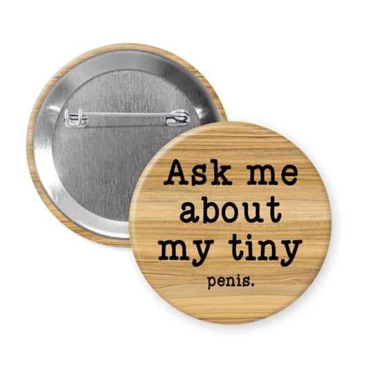 Button badge with text 'Ask me about my tiny penis' on a wooden background