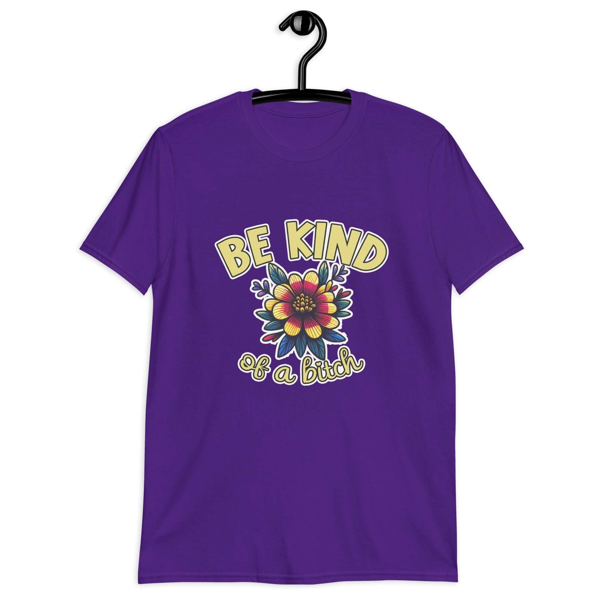 Purple t-shirt with an image of a flower and the words Be kind above the flower in yellow bold block font. The words Of a bitch are smaller in script font under the flower. The design is printed on the front of the shirt.