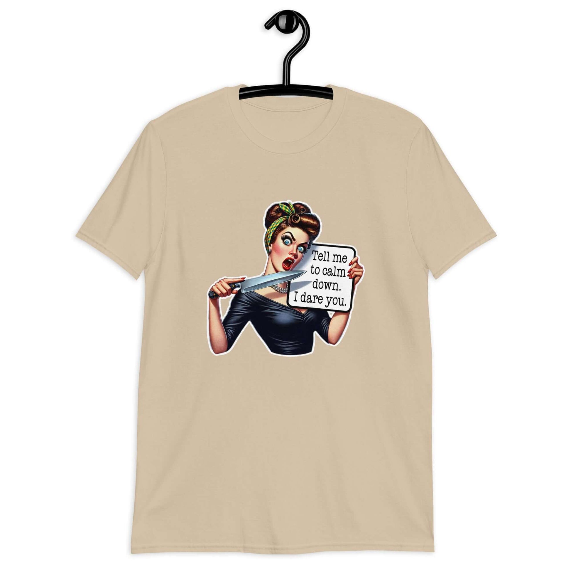 Sand color t-shirt displayed on a hanger. The t-shirt has a graphic of an angry looking retro woman holding a knife and a sign. The sign says Tell me to calm down I dare you. The graphic is printed on the front of the shirt.