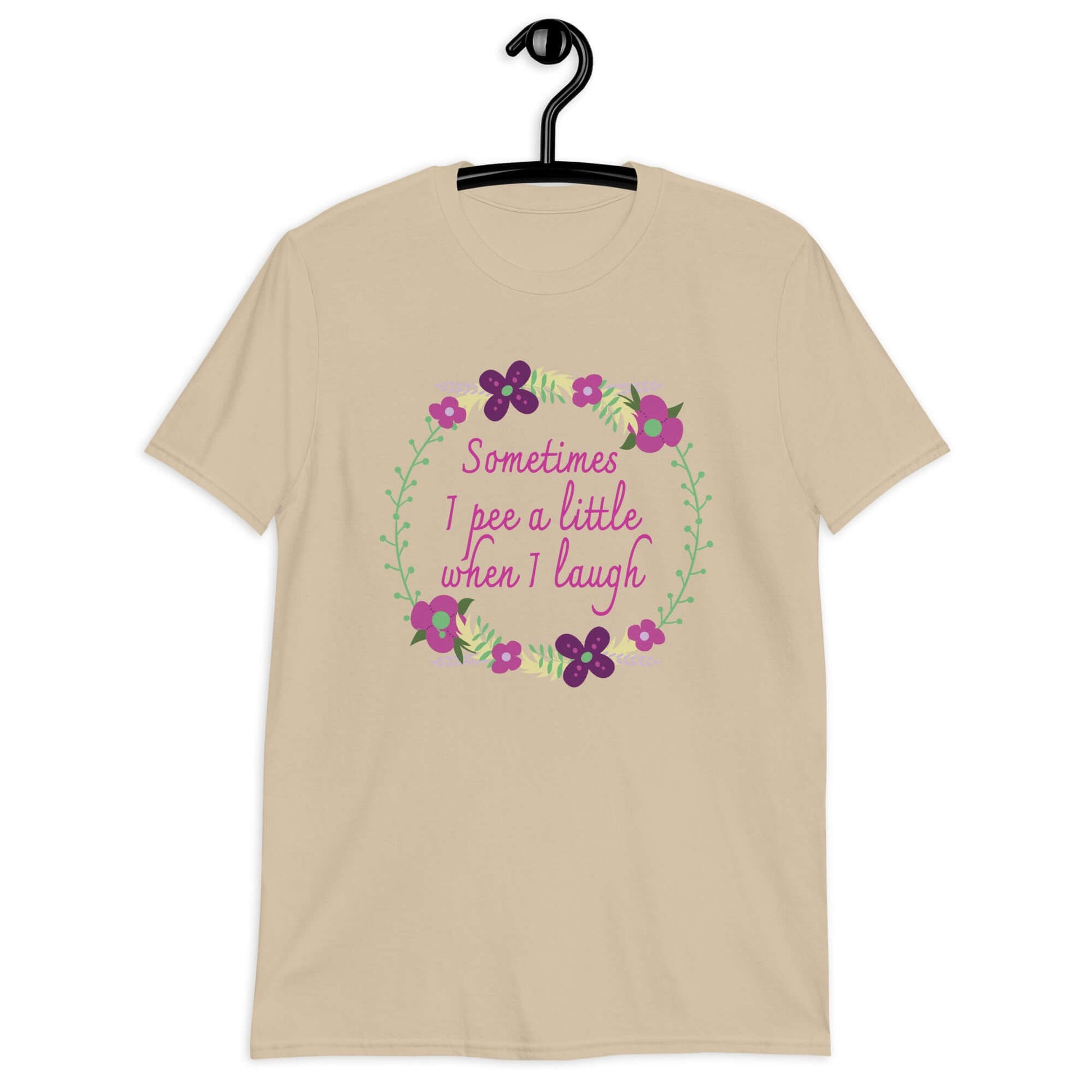 Sand color t-shirt that has a minimalistic style line drawing floral wreath with the phrase Sometimes I pee a little when I laugh printed in the center of the wreath. The graphics are printed on the front of the shirt.