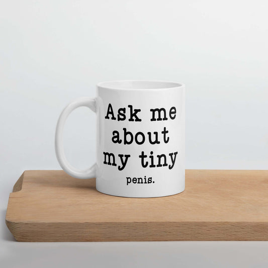 11 ounce white ceramic mug with the phrase Ask me about my tiny penis printed on both sides. The word tiny is much smaller than the rest of the text.