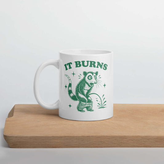White ceramic mug with an image of an opossum peeing. The words It burns are printed above.