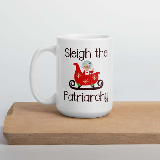 White ceramic mug with graphic of Mrs. Claus sitting in a sleigh and the words Sleigh the patriarchy printed on both sides.