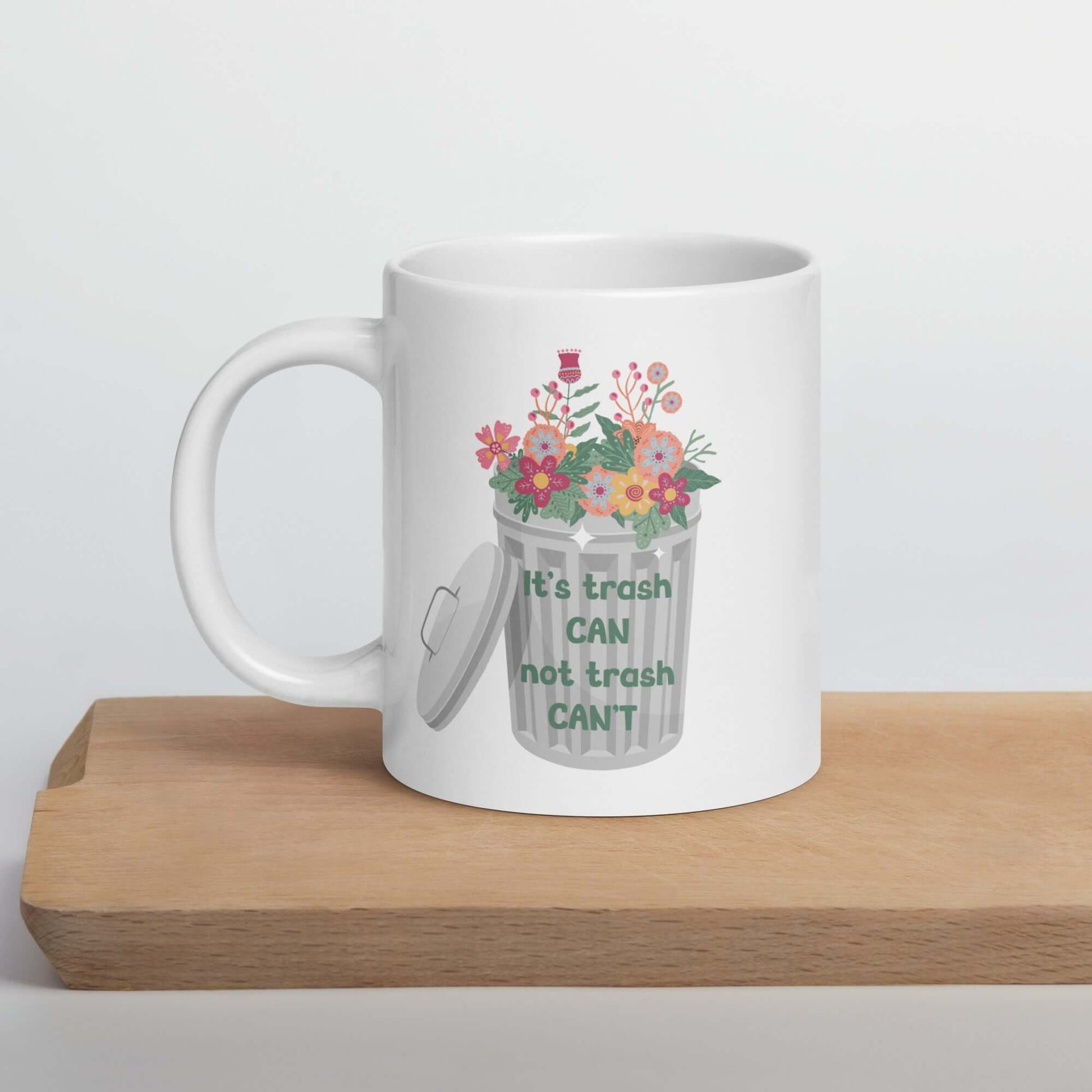 White ceramic coffee mug with graphic of trash can with flowers growing in it. The phrase It's trash can not trash can't is printed on the trash can. The graphics are printed on both sides of the mug.