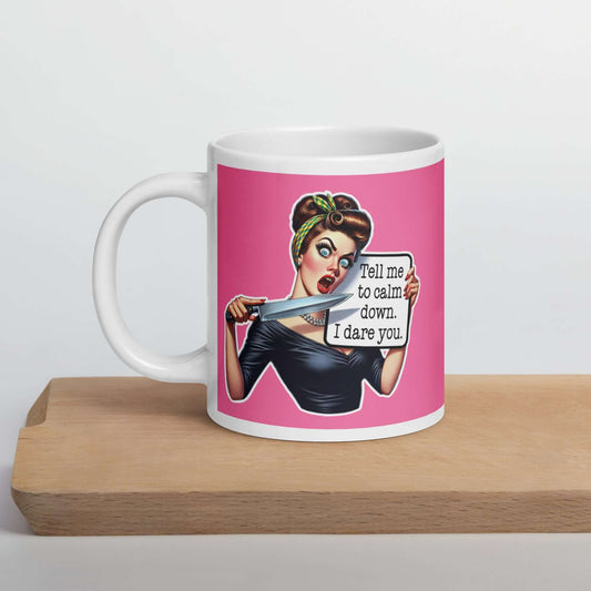 White ceramic coffee mug with pink color wrapped around. The mug has a graphic of an angry looking retro woman holding a knife and a sign. The sign says Tell me to calm down I dare you. The graphic is printed on both sides of the mug.