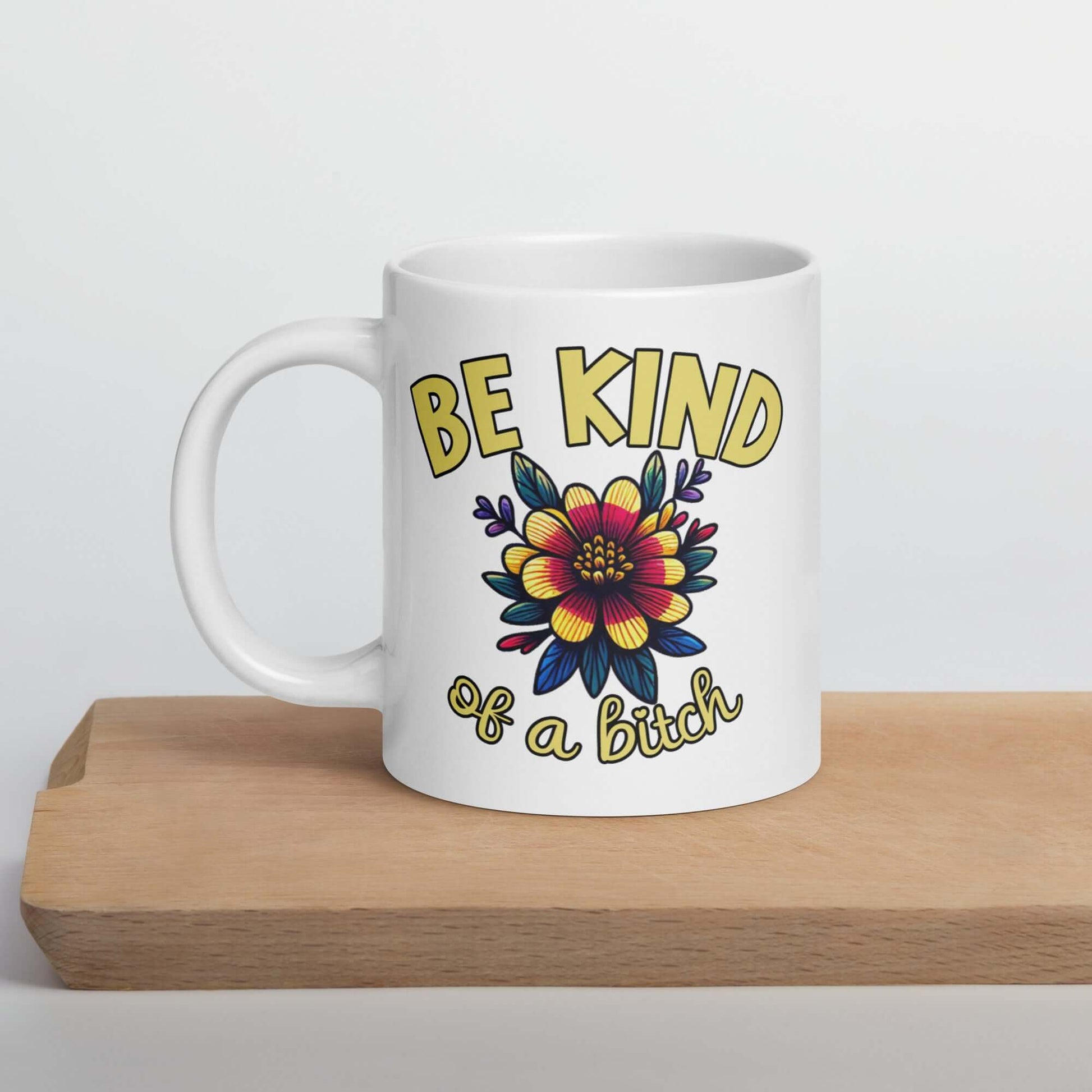 White ceramic coffee mug with an image of a flower and the words Be kind above the flower in bold block font. The words Of a bitch are smaller in script font under the flower. The design is printed on both sides of the mug.