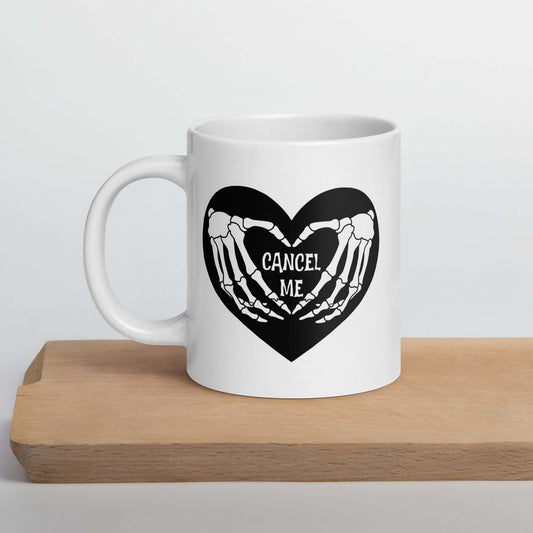 White ceramic mug with a black heart and skeleton hands making a heart shape. The words Cancel Me are inside of the heart. The graphics are printed on both sides of the mug.