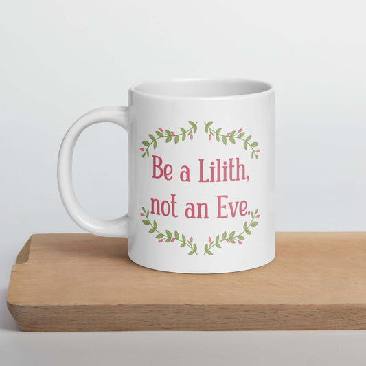 White ceramic mug with the phrase Be a Lilith, not an Eve printed on both sides. The text is pink and has a green floral vine graphic framing the text. 