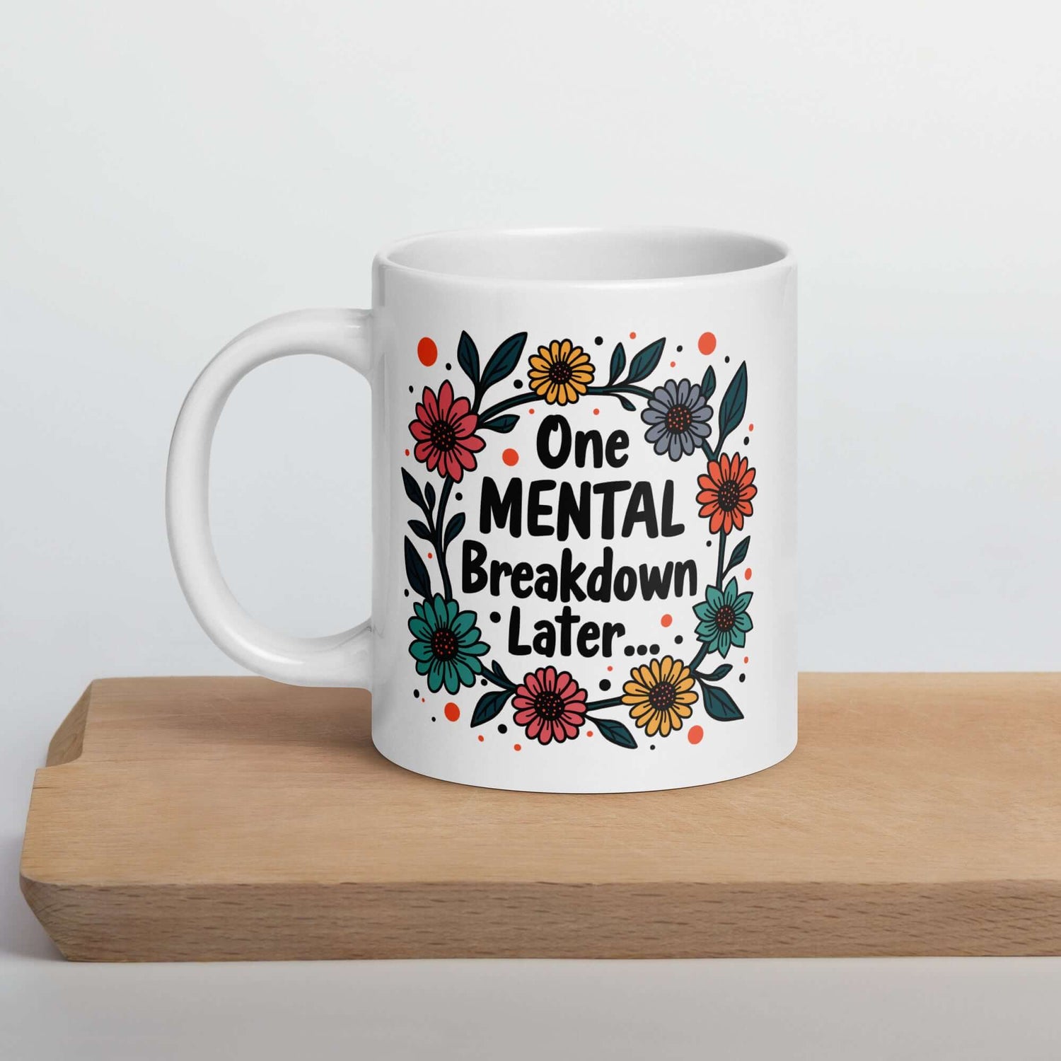 White ceramic mug with a multi color floral wreath design printed on it. The words One mental breakdown later are printed in the center of the wreath. The graphics are printed on both sides of the mug.