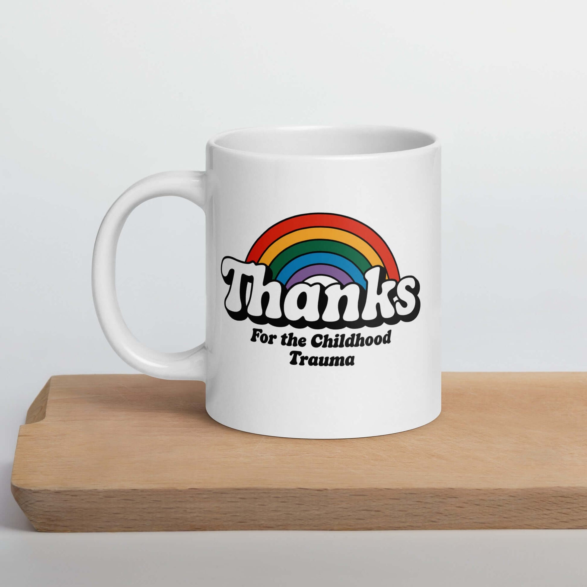 White ceramic mug with an image of a rainbow and the words Thanks for the childhood trauma printed on both sides of the mug. The word Thanks is large and the rest of the words are smaller under.