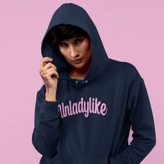 Woman wearing navy blue hoodie sweatshirt with the hood up partially covering her face. The hoodie has the word Unladylike printed on the front in pink.