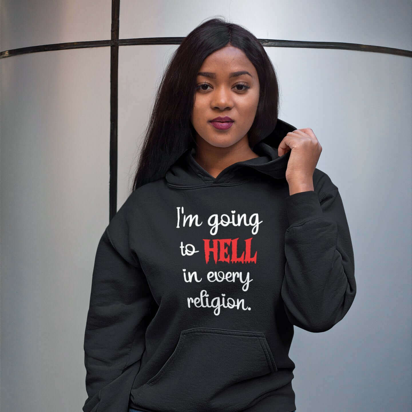 Woman wearing black hoodie sweatshirt with the phrase I'm going to hell in every religion printed on the front. The word hell is printed in red. The rest of the text is white.