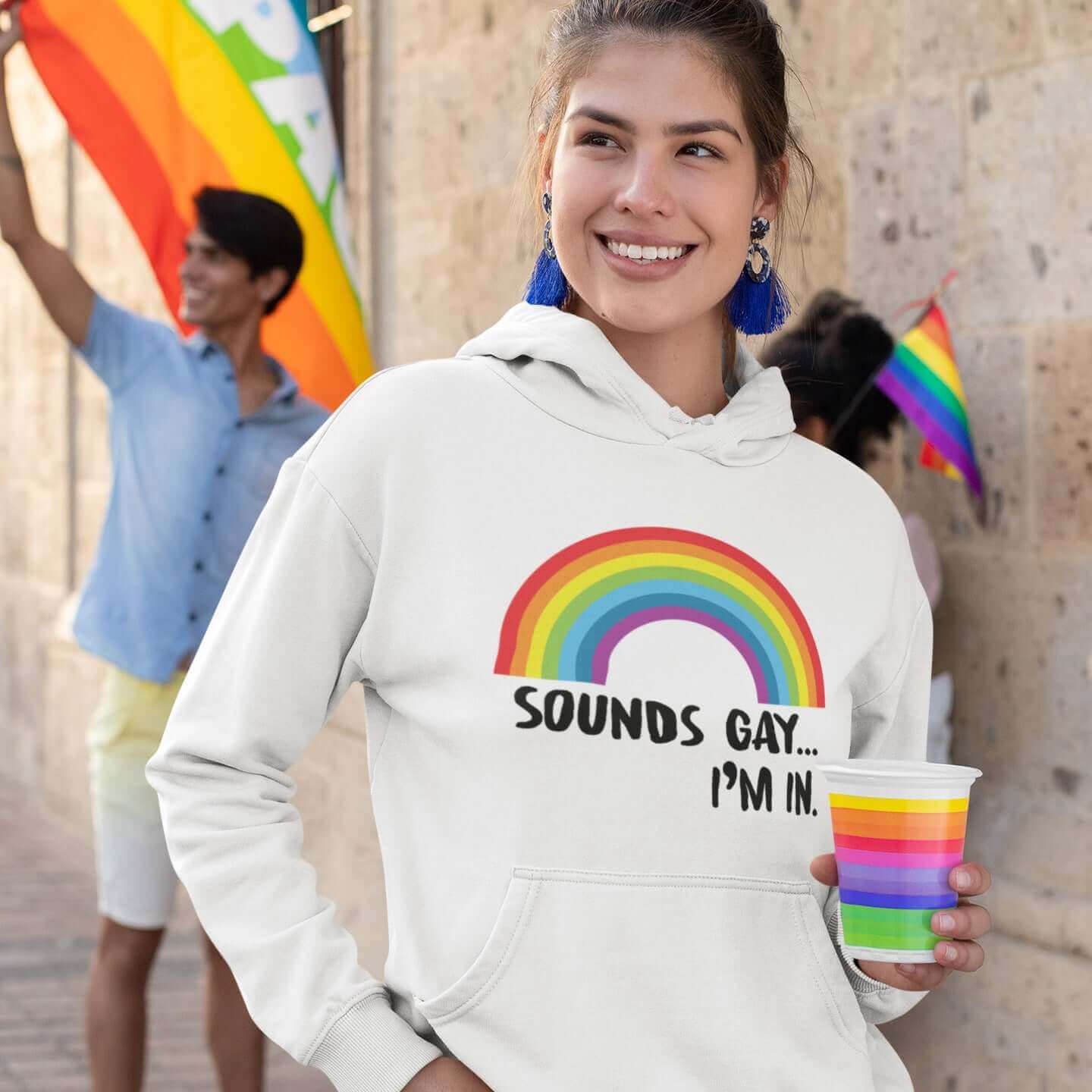 Woman at a pride event wearing a white hoodie sweatshirt that has an image of a rainbow and the phrase Sounds gay, I'm in printed on the front