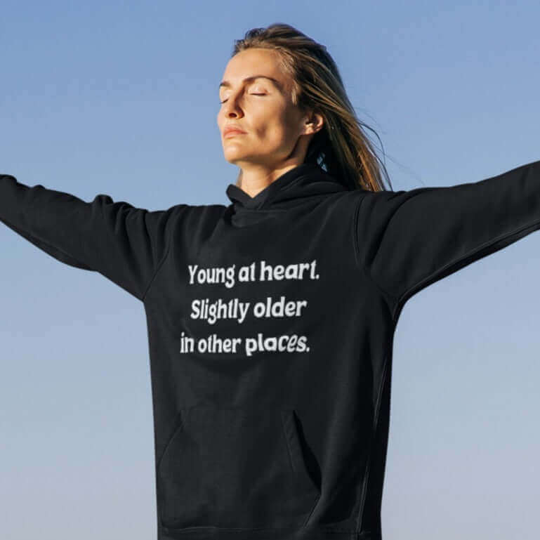 Woman standing with arms outstretched wearing a black hoodie sweatshirt with the words Young at heart, slightly older in other places printed on the front.