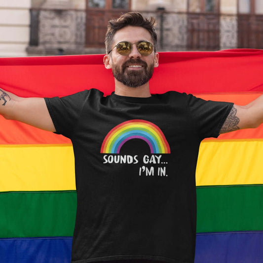 Man at a pride event wearing a black t-shirt that has an image of a rainbow and the phrase Sounds gay, I'm in printed on the front