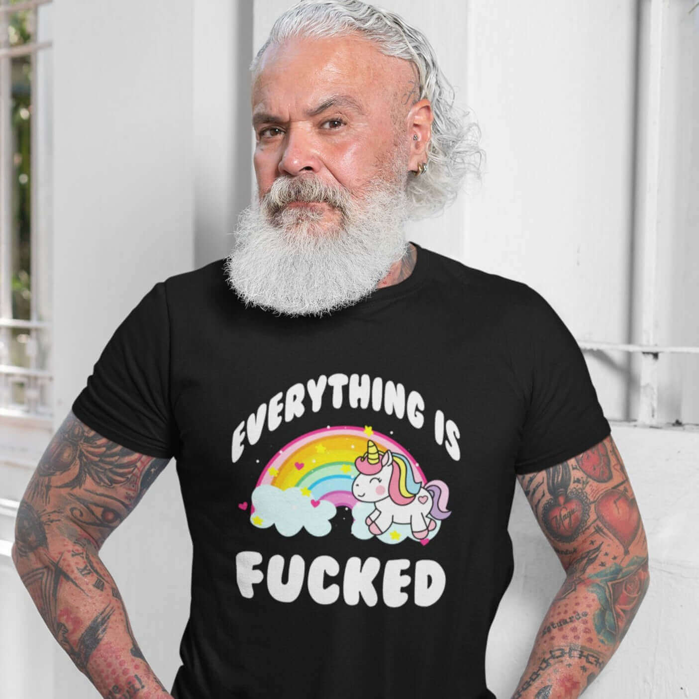 Tattooed older man with a beard wearing a black t-shirt with a graphic of a kawaii style unicorn and a pastel rainbow with the words Everything is fucked printed on the front.