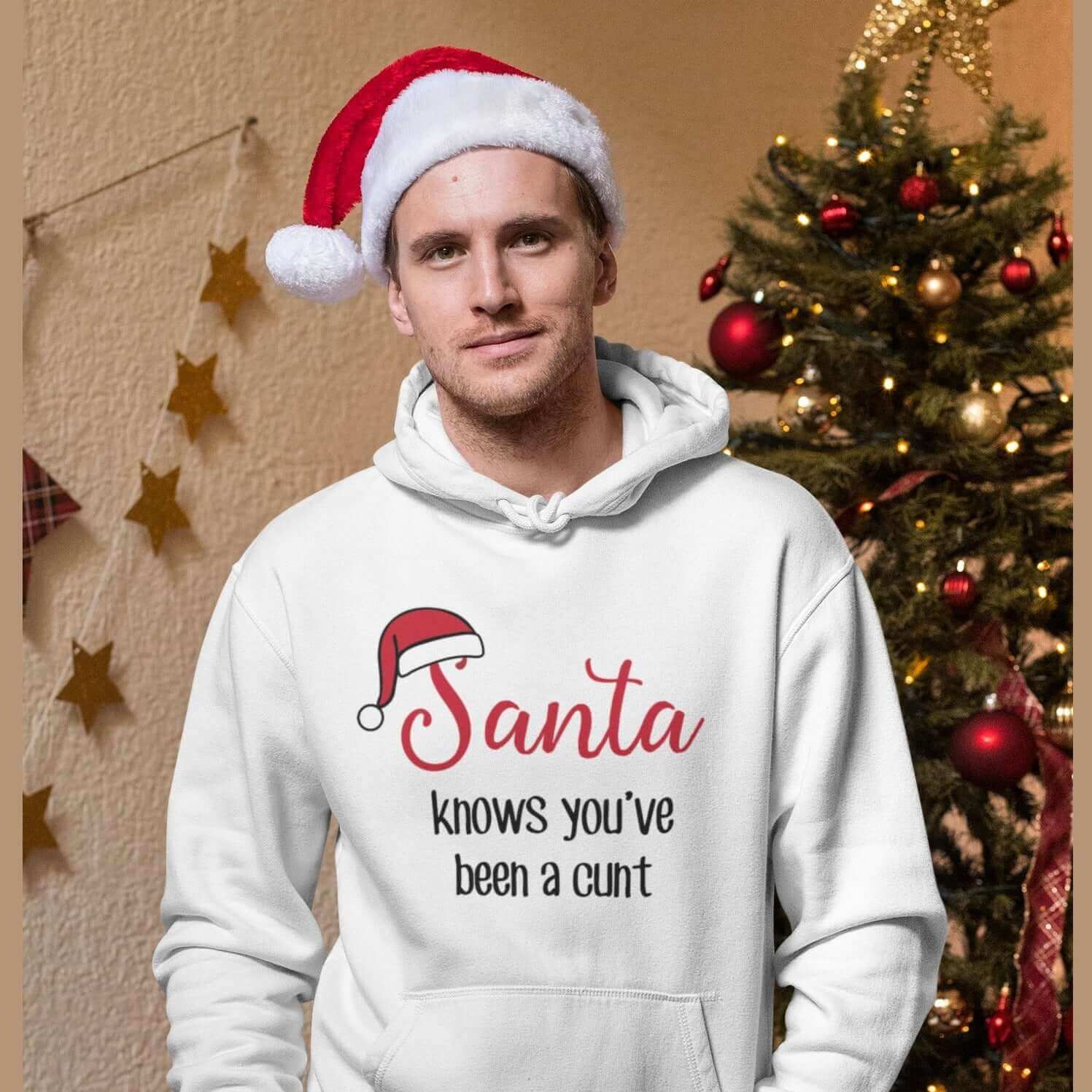 Man wearing santa hat standing next to Christmas tree. He is wearing a white hoodie sweatshirt with the words Santa knows you've been a cunt printed on the front. There is a Santa hat on the s in Santa.