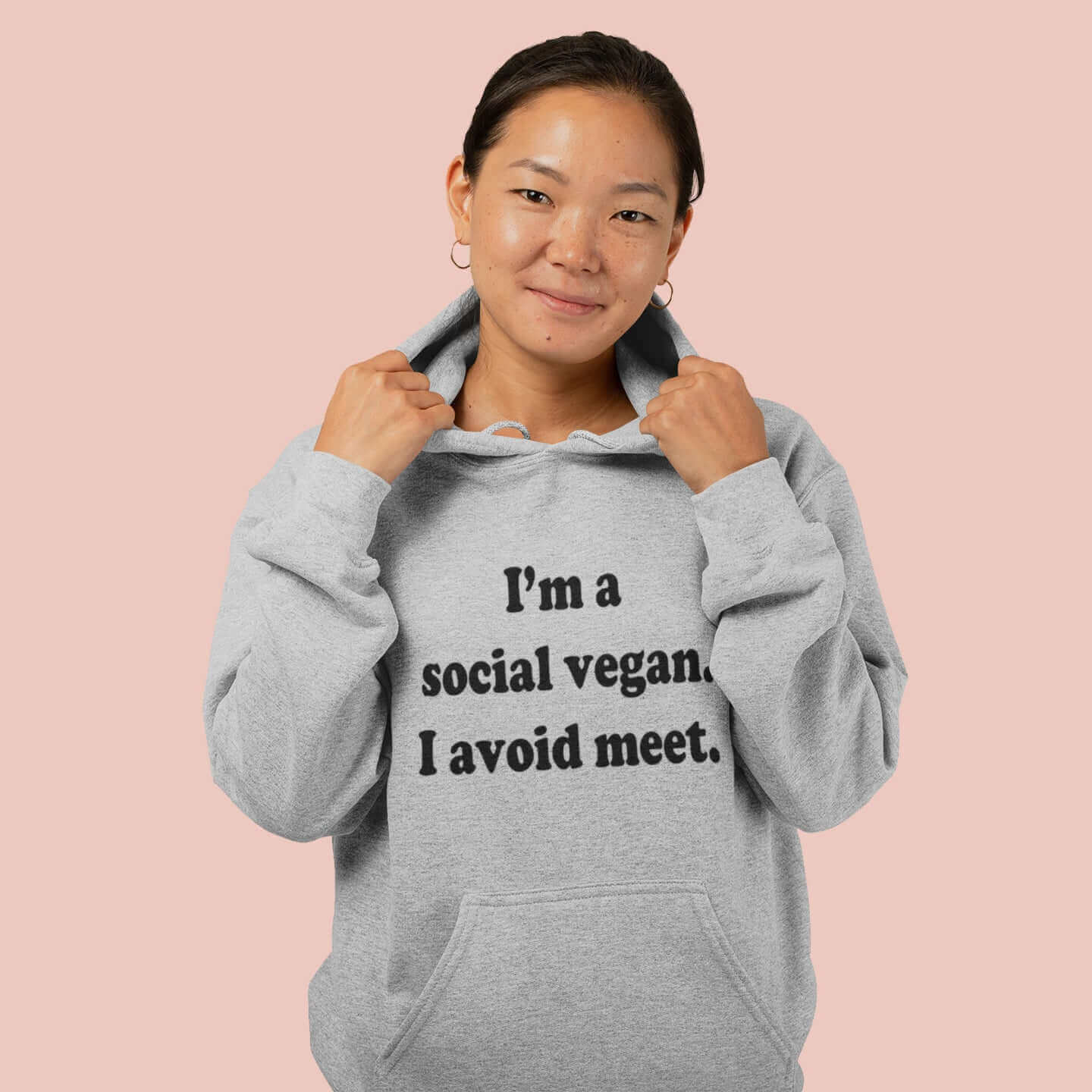 Woman wearing light grey hoodie sweatshirt with the pun phrase I'm a social vegan, I avoid meet printed on the front.