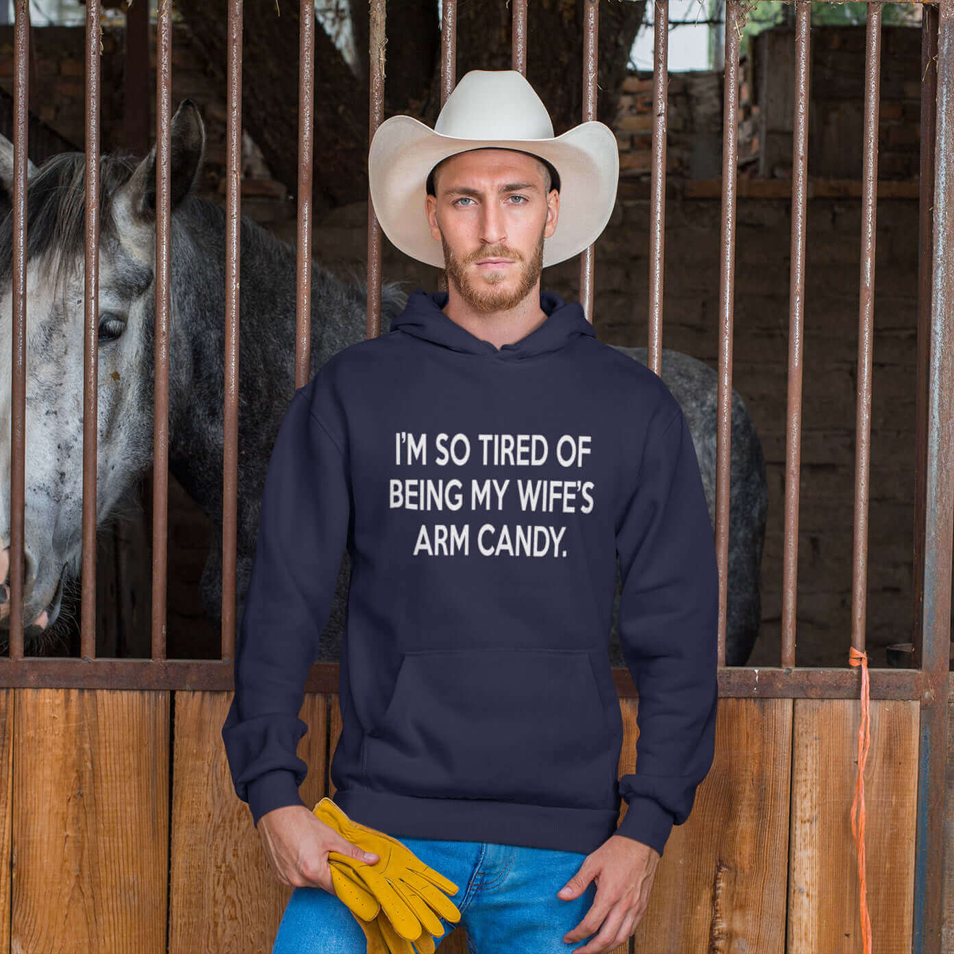Man wearing a cowboy hat standing in a stable. He is wearing a navy blue hoodie sweatshirt with the funny phrase I'm so tired of being my wife's arm candy printed on the front.