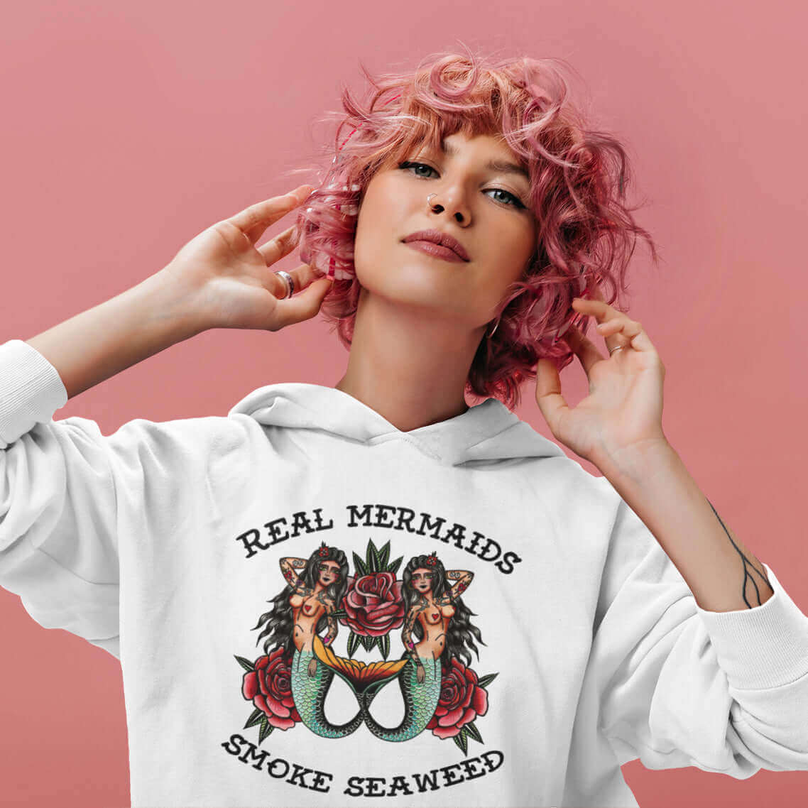 Woman with pink curly hair wearing a white hooded sweatshirt with image of 2 mermaids and the words Real mermaids smoke seaweed printed on the front.