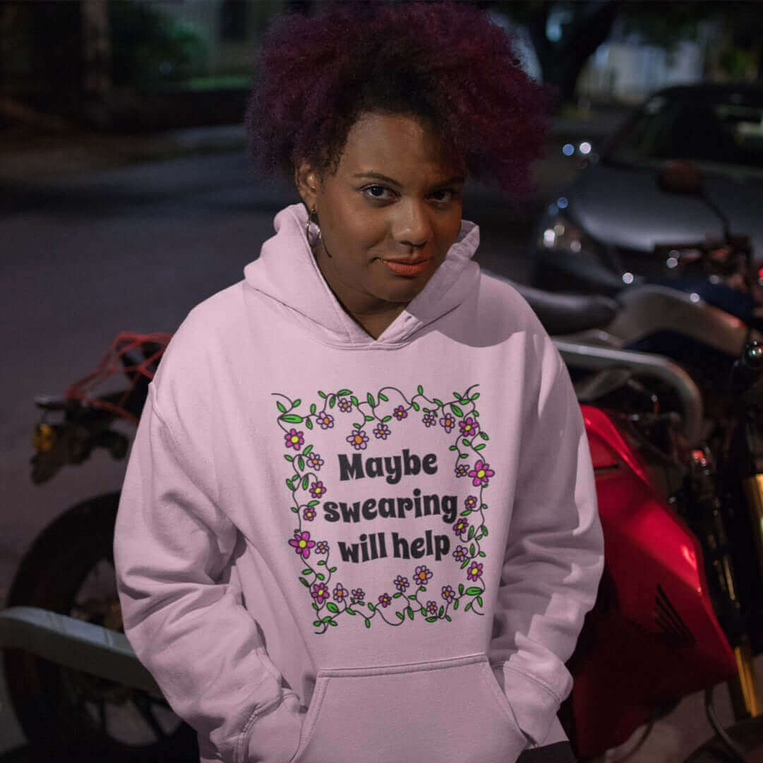 Woman wearing light pink hoodie sweatshirt with a floral graphic and the phrase Maybe swearing will help printed on the front.
