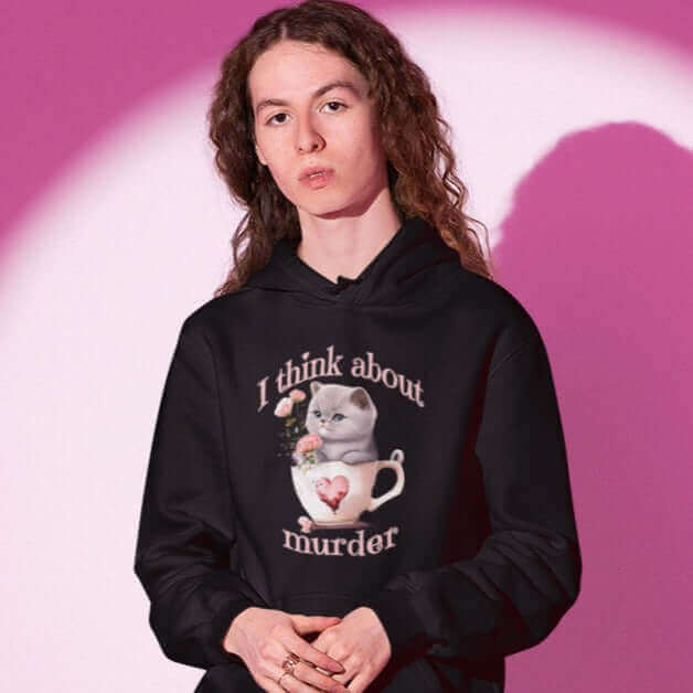 Man wearing black hooded sweatshirt that says I think about murder with image of cute fluffy kitten sitting in a teacup printed on the front.