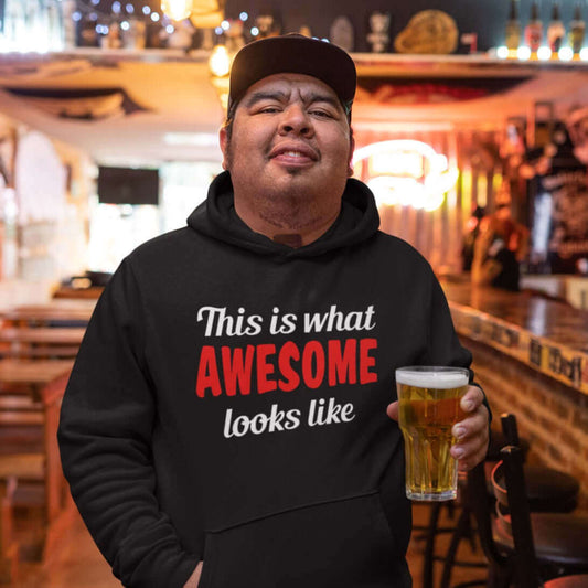 Man holding a beer wearing a black hoodie sweatshirt with the words This is what awesome looks like printed on the front. The word awesome is bright red. The rest of the text is white.