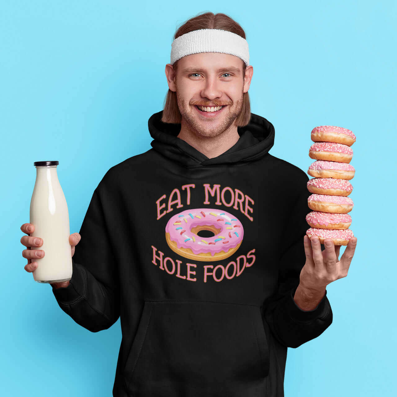 Man holding a stack of donuts and a bottle of milk. He is wearing a black hoodie sweatshirt with an image of a donut with pink icing and sprinkles and the words Eat more hole foods printed on the front.