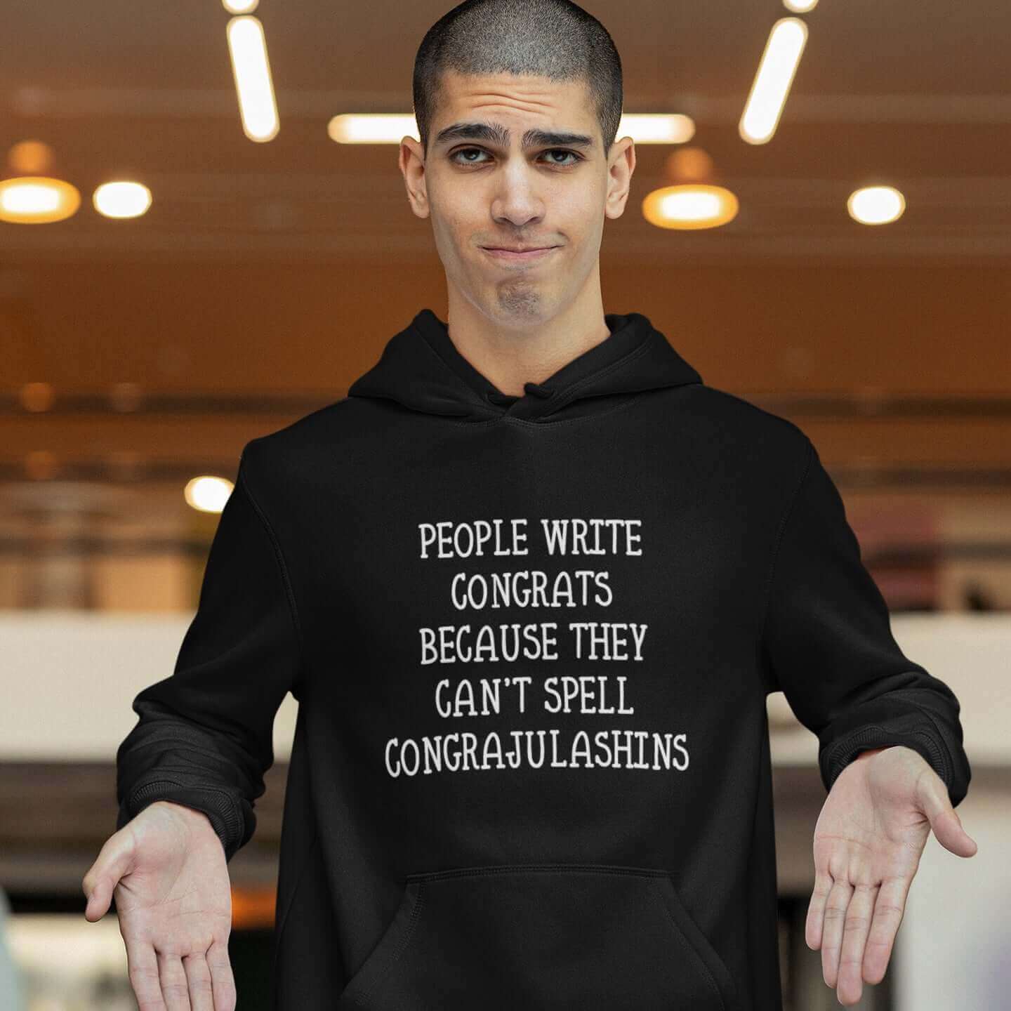 Man wearing black hooded sweatshirt with the words People write congrats because they can't spell congratulations printed on the front. The word congratulations is intentionally misspelled.