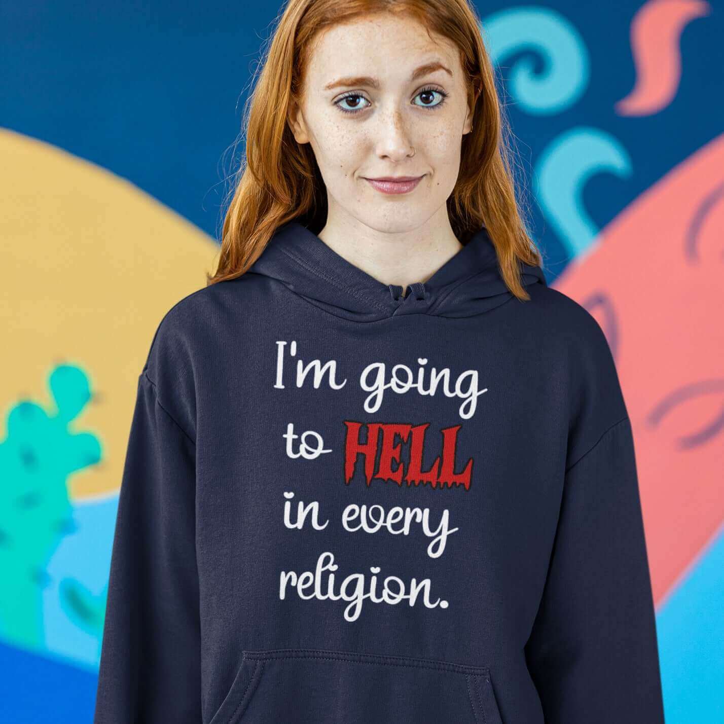 Woman wearing a navy blue hoodie sweatshirt with the phrase I'm going to hell in every religion printed on the front. The word hell is printed in red. The rest of the text is white.