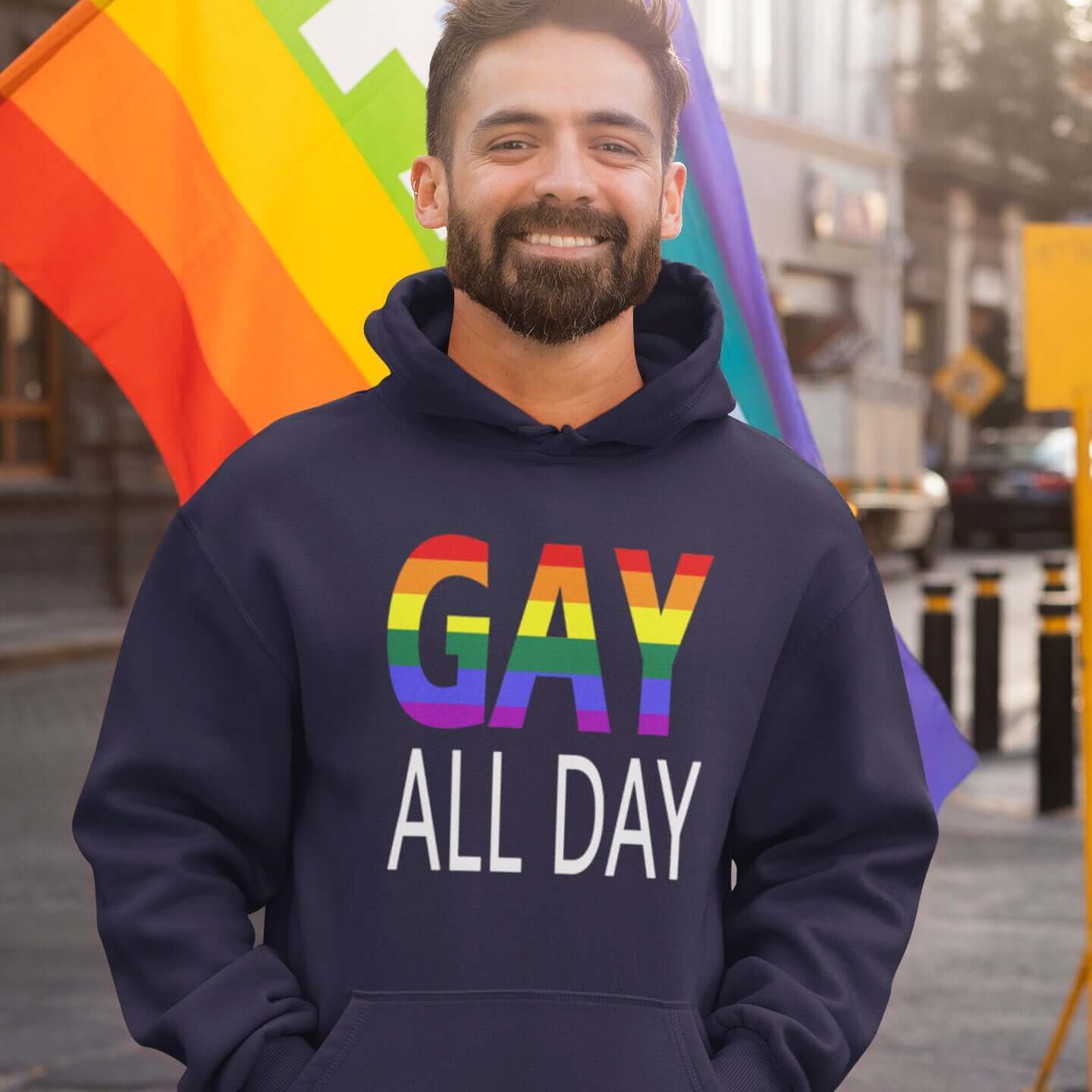 Man at a pride event wearing a navy blue hoodie sweatshirt with the words Gay all day printed on the front. The word Gay is in rainbow stripe font. 