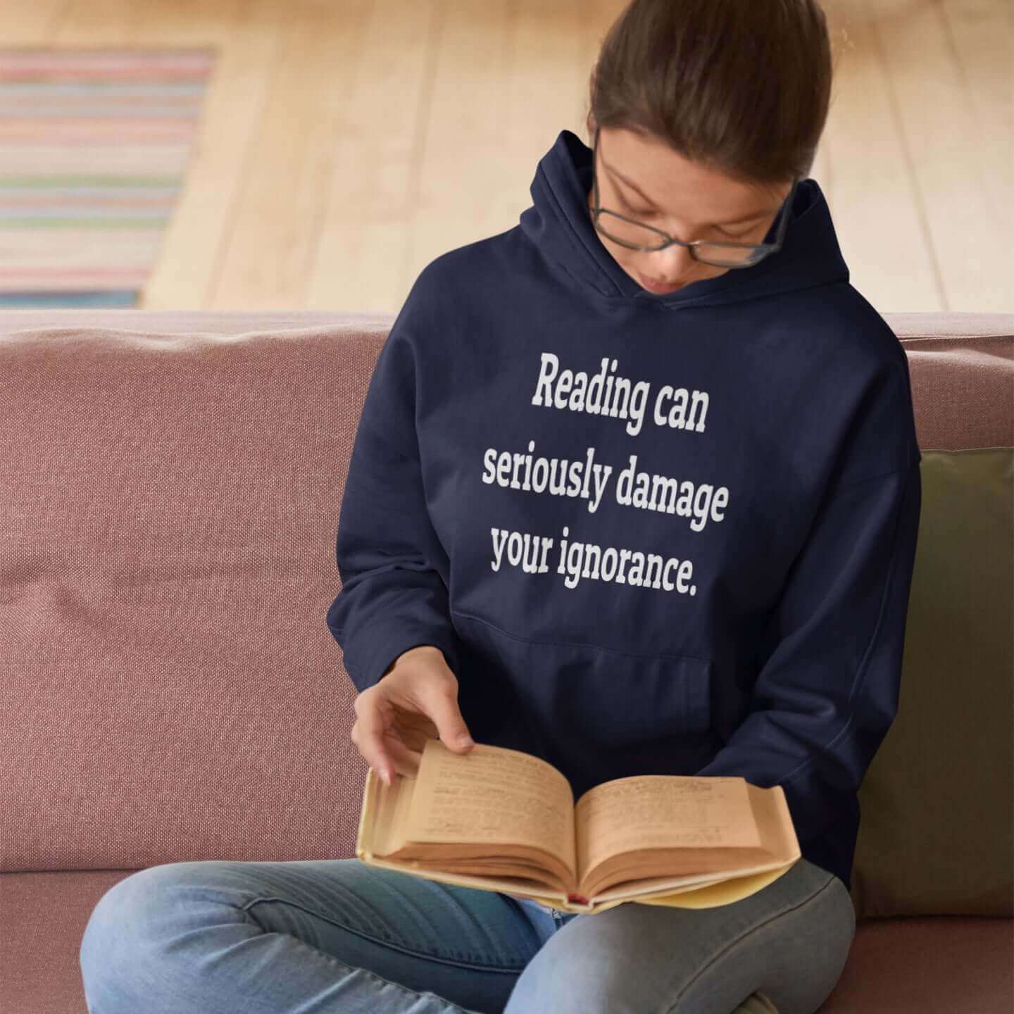 Woman reading a book while wearing a navy hoodie sweatshirt with the phrase Reading can seriously damage your ignorance printed on the front.