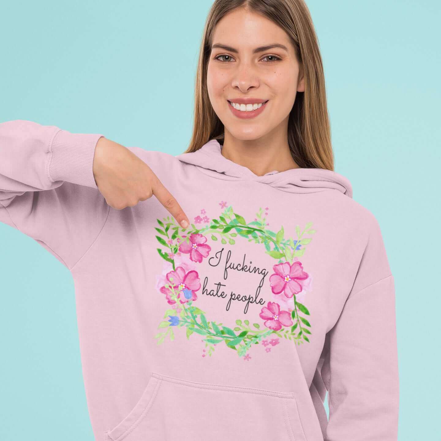 Woman wearing light pink hooded sweatshirt with pink and green floral wreath image and the words I fucking hate people printed on the front.