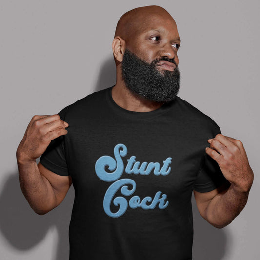 Bearded man gesturing to the front of his T-shirt. He is wearing a black t-shirt with the words Stunt Cock printed on the front in blue.