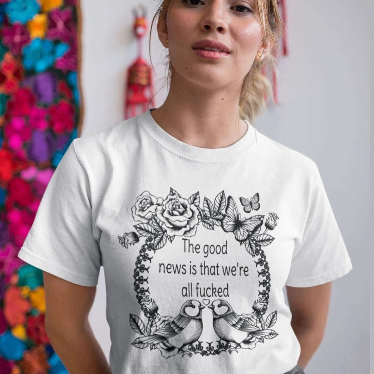 Woman wearing a white t-shirt that has an image of a line drawing wreath with butterflies, sparrows and roses. The phrase The good news is that we're all fucked printed inside of the wreath. The graphics are printed on the front of the shirt.