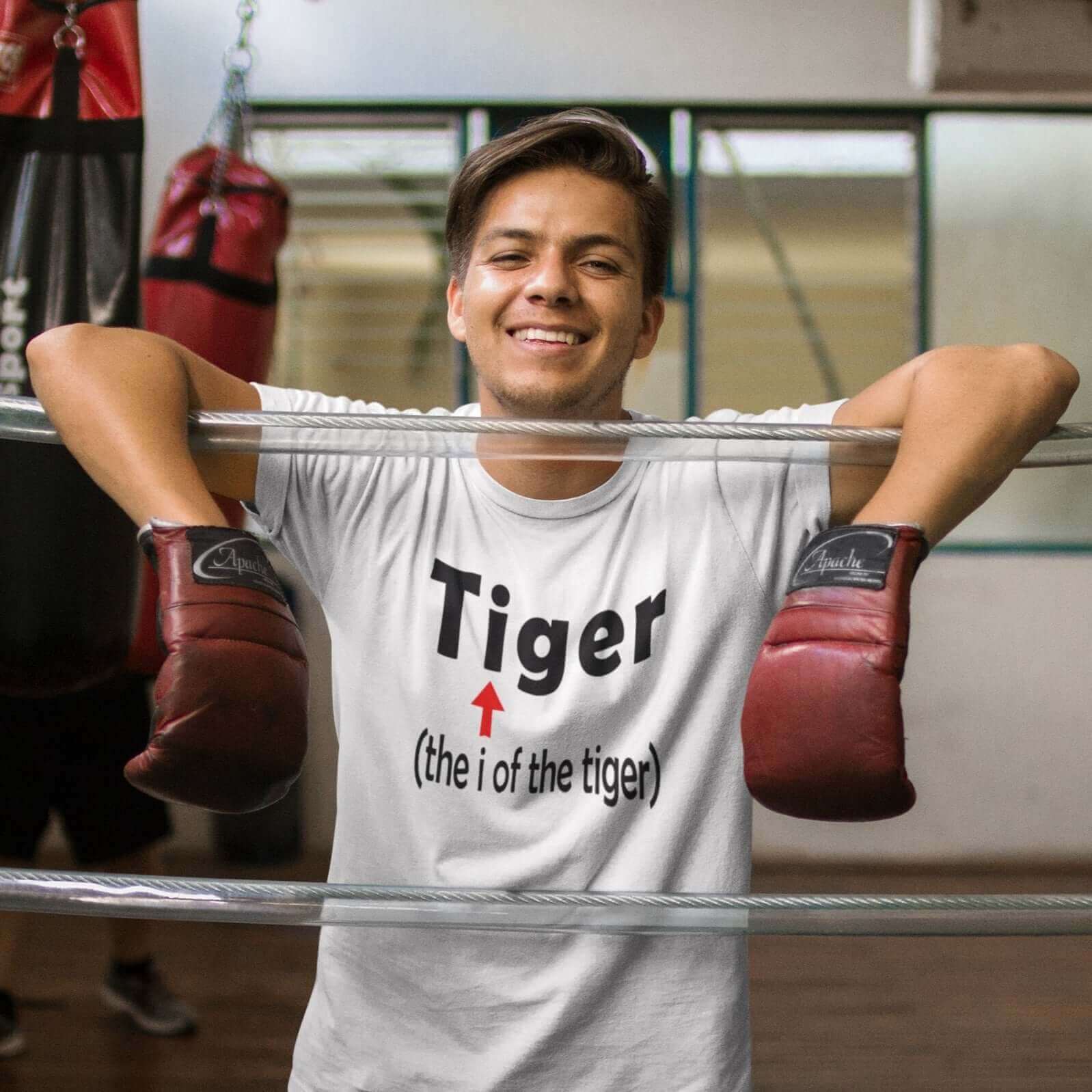 Man in a boxing ring wearing boxing gloves and a white t-shirt. The t-shirt has the word Tiger and in smaller letters it says the I of the tiger with an arrow pointing to the letting I in the words tiger. The graphics are printed on the front of the shirt
