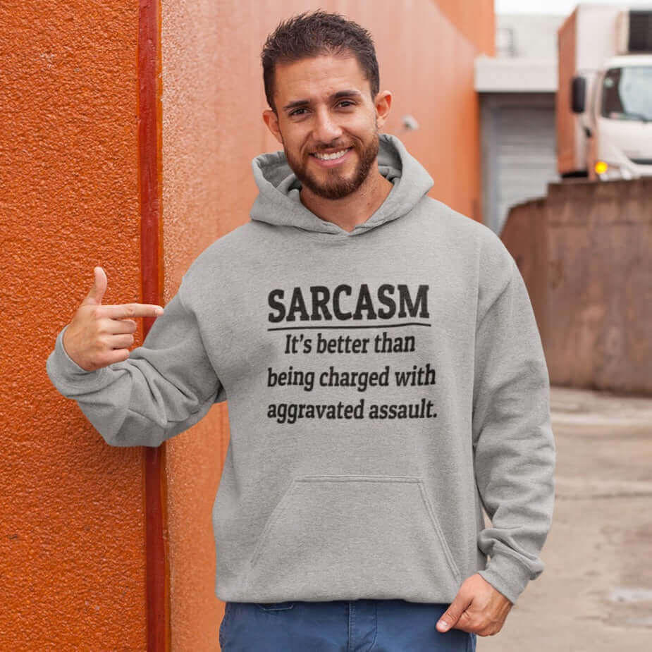 Man wearing a light grey hoodie sweatshirt pointing to the phrase Sarcasm, it's better than being charged with aggravated assault printed on the front of the hoodie.