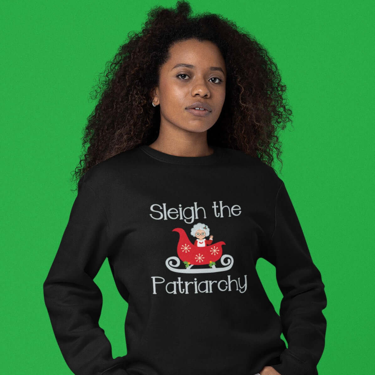 Woman in festive setting wearing a black crewneck sweatshirt with graphic of Mrs. Claus sitting in a sleigh and the words Sleigh the patriarchy printed on the front.