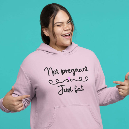 Winking woman pointing at the front of her sweatshirt. She is wearing a light pink hoodie sweatshirt with the words Not pregnant just fat printed on the front with a heart.