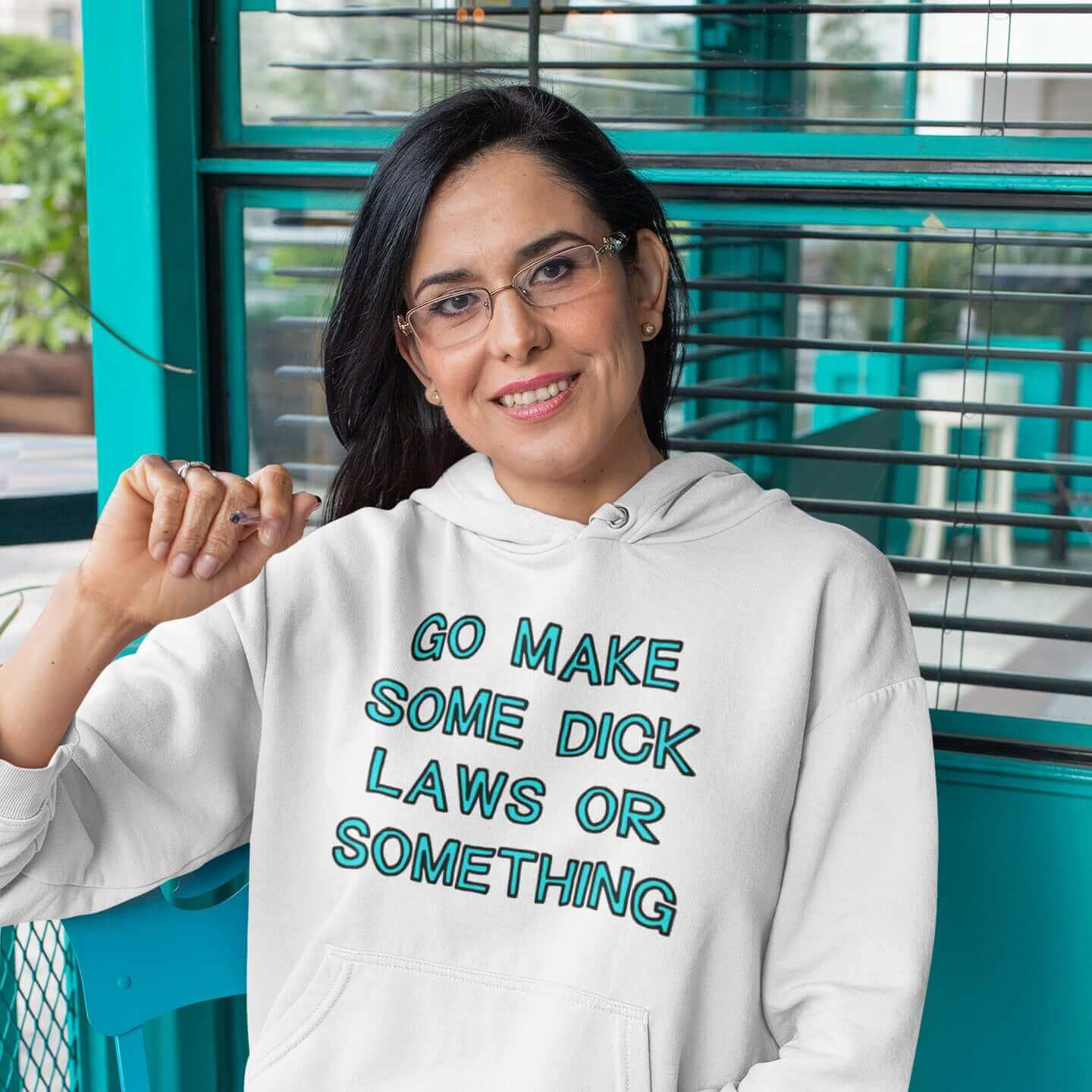 Woman sitting at a cafe table wearing a white hoodie sweatshirt with the words Go make some dick laws or something printed on the front. The text is turquoise with black outline.