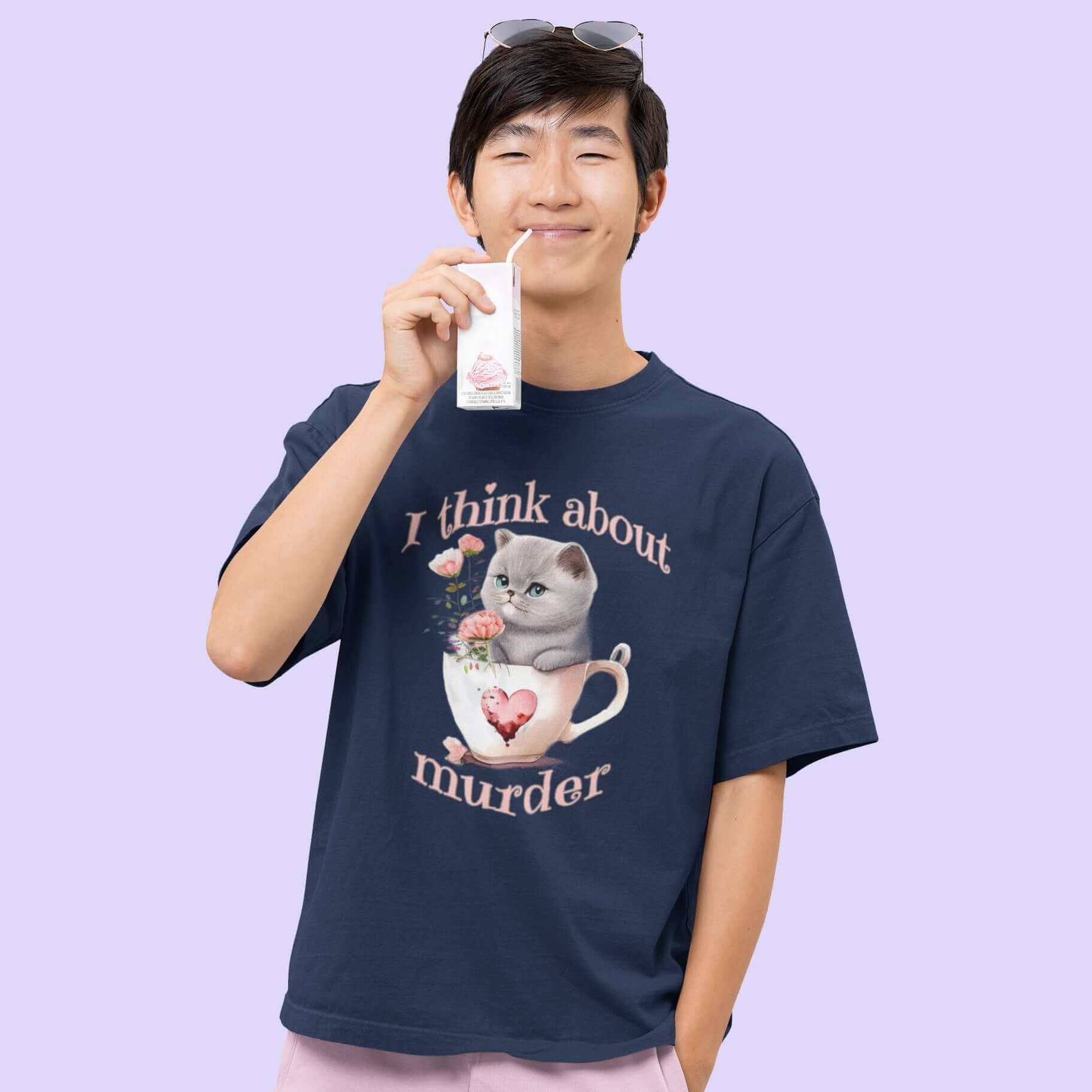 Man sipping juice box wearing a navy blue t-shirt that says I think about murder with image of cute fluffy kitten sitting in a teacup printed on the front.