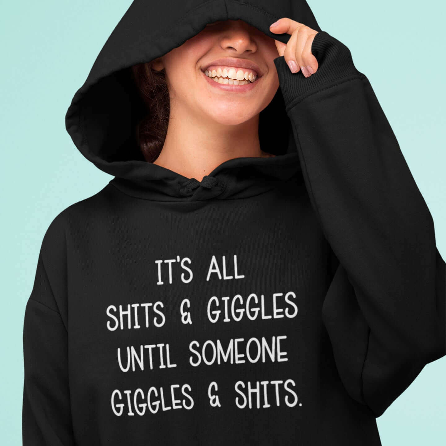 Smiling woman wearing a black hoodie sweatshirt with the words It's all shits & giggles until someone giggles & shits printed on the front.