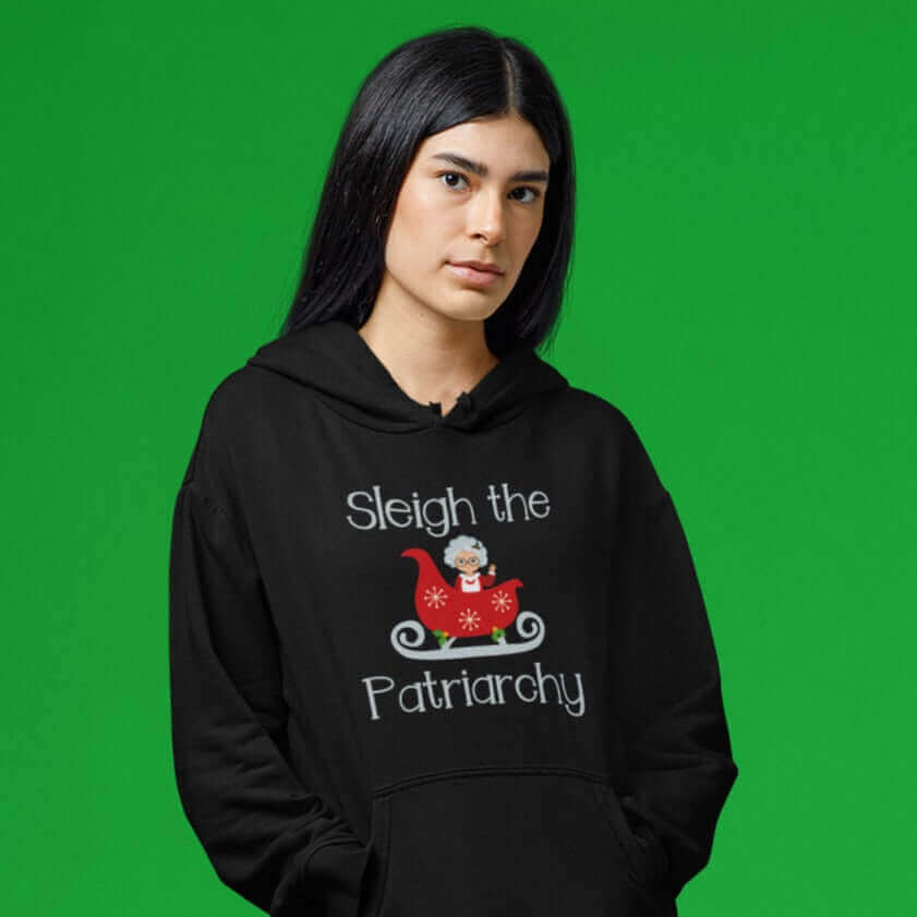 Woman in festive setting wearing a black hooded sweatshirt with graphic of Mrs. Claus sitting in a sleigh and the words Sleigh the patriarchy printed on the front.