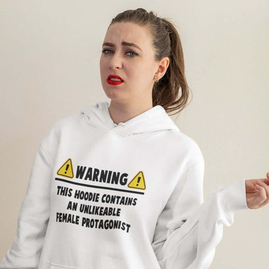 Woman with annoyed look on her face wearing a white hoodie sweatshirt with the words Warning this hoodie contains an unlikable female protagonist printed on the front.