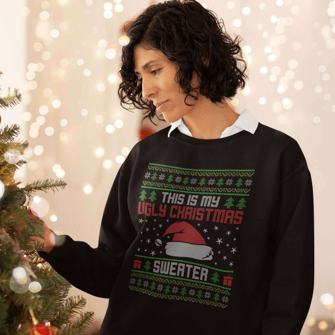 Woman standing next to Christmas tree wearing a black crewneck sweatshirt with the words This is my ugly Christmas sweater printed on the front. 