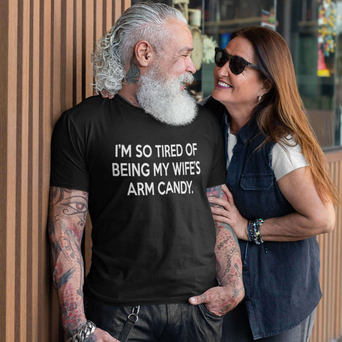 Tattooed bearded man wearing a black t-shirt with the funny phrase I'm so tired of being my wife's arm candy printed on the front.