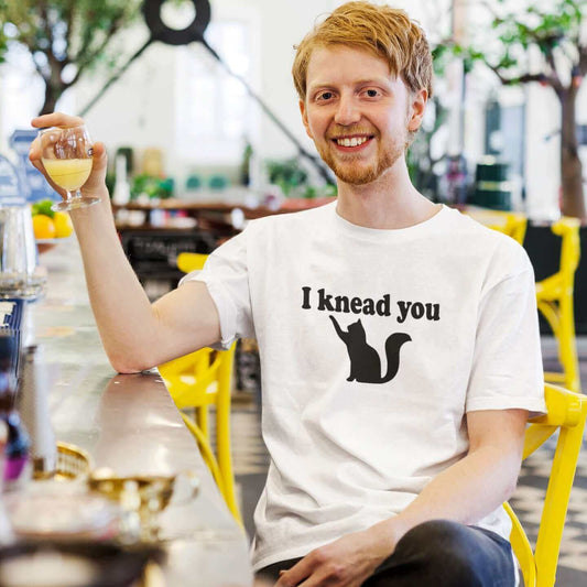 Man wearing a white t-shirt that has a pun image of a silhouette of a cat and the words I knead you printed on the front.