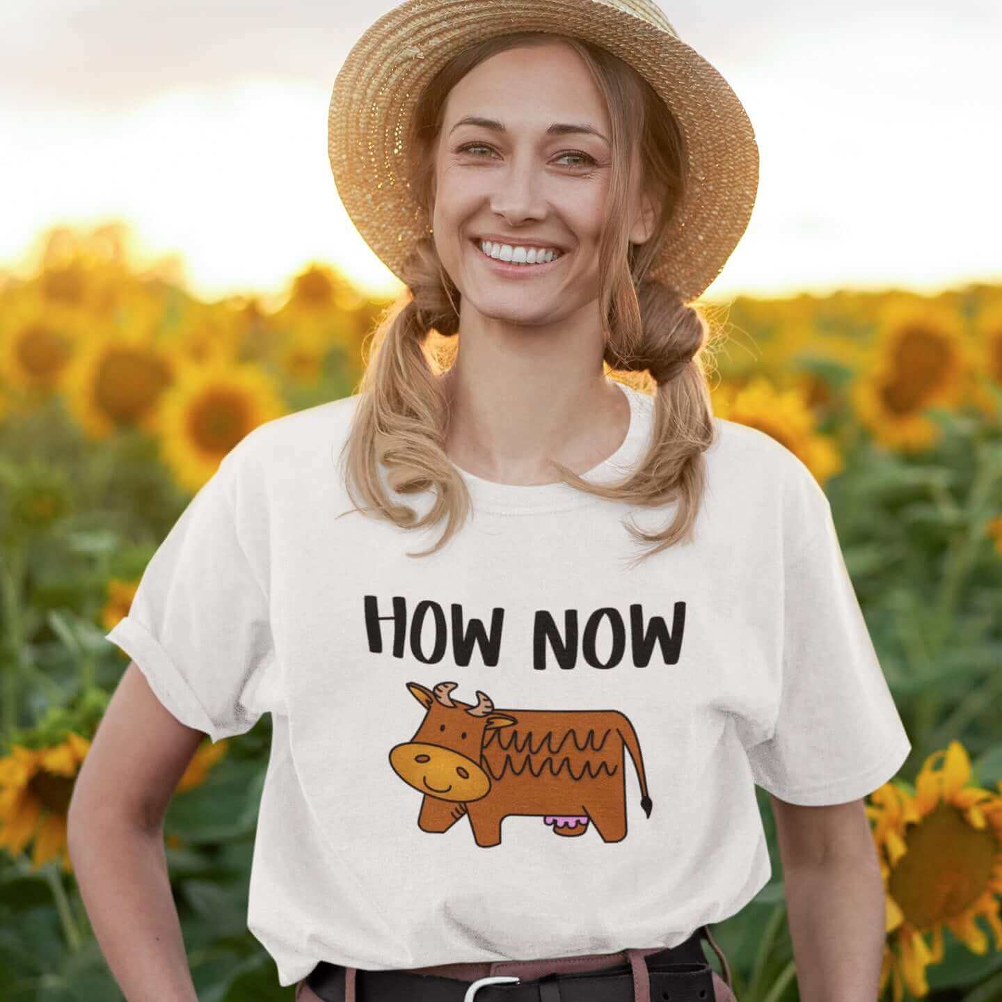 Woman standing in a field of sunflowers wearing a white t-shirt with the words How now and an image of a brown cow printed on the front.